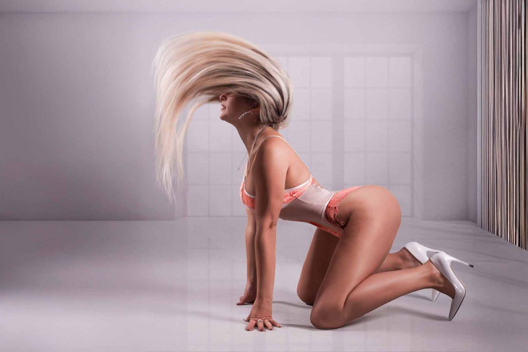 ine Frau mit langem, blondem Haar, das dynamisch nach oben fliegt, kniet auf dem Boden. Sie trägt einen hellen Body mit Spitzen- oder Transparenzdetails und weiße High Heels.