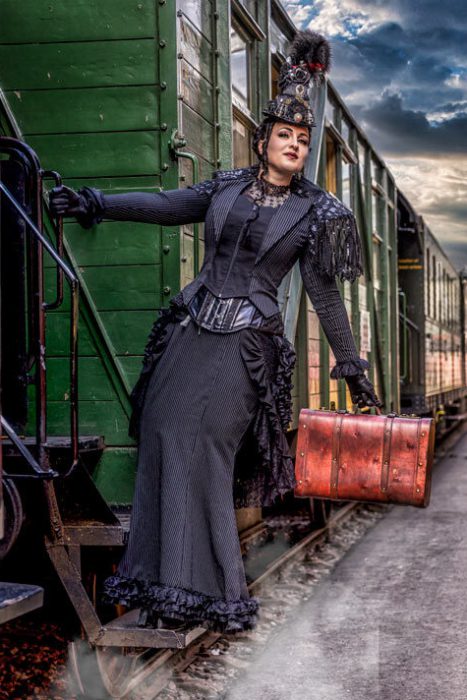 Eine Frau in einem schwarzen, gestreiften, viktorianisch/Steampunk-inspirierten Kleid mit Korsett und Hut, die mit einem braunen Vintage-Koffer die Stufen eines grünen Eisenbahnwaggons betritt.
