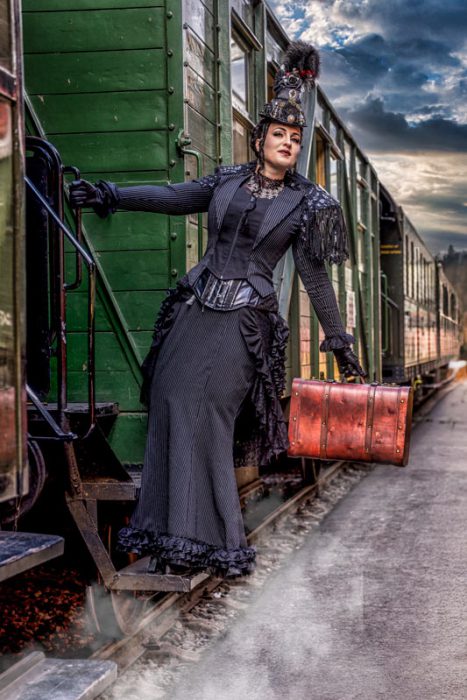 Eine Frau in einem schwarzen, gestreiften, viktorianisch/Steampunk-inspirierten Kleid mit Korsett und Hut, die mit einem braunen Vintage-Koffer die Stufen eines grünen Eisenbahnwaggons betritt.