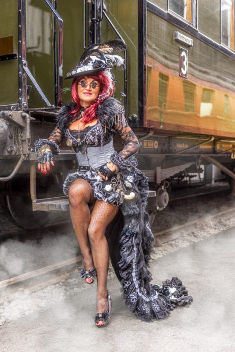 Eine Frau mit leuchtend rotem Haar in einem aufwendigen, schwarzen Steampunk-Kostüm mit Korsett und kurzem Rock, sitzt lächelnd auf der Trittstufe eines historischen, grünen Zugwaggons.