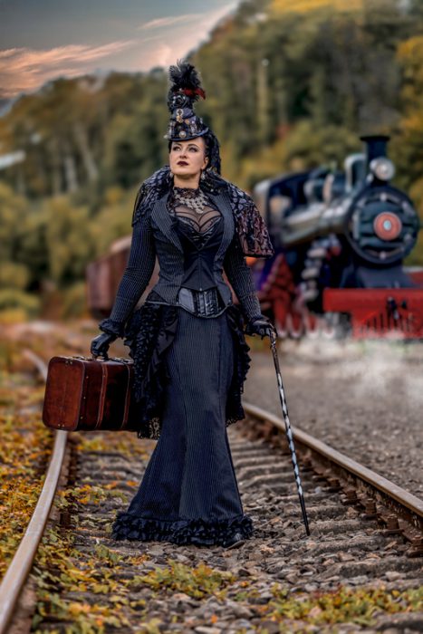 Eine elegant gekleidete Frau in einem schwarzen, gestreiften Steampunk-Kleid mit Korsett und Hut, die mit einem Vintage-Koffer und einem Stock auf Bahngleisen steht. Im Hintergrund ist eine Dampflokomotive unscharf zu sehen.