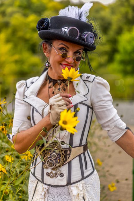 Eine Frau mittleren Alters in einem weißen, korsettartigen Steampunk-Outfit mit schwarzen Akzenten, einem Zylinder mit Feder und Schutzbrille, hält eine gelbe Blume in einem Feld.