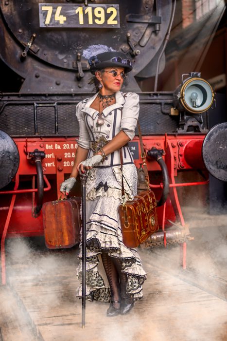 Eine Frau in einem weißen Steampunk-Korsett-Outfit mit Rüschenrock, Hut und Nickelbrille steht mit einem kleinen Koffer, einer Umhängetasche und einem Spazierstock vor der roten Pufferbohle und dem schwarzen Kessel einer historischen Dampflokomotive.