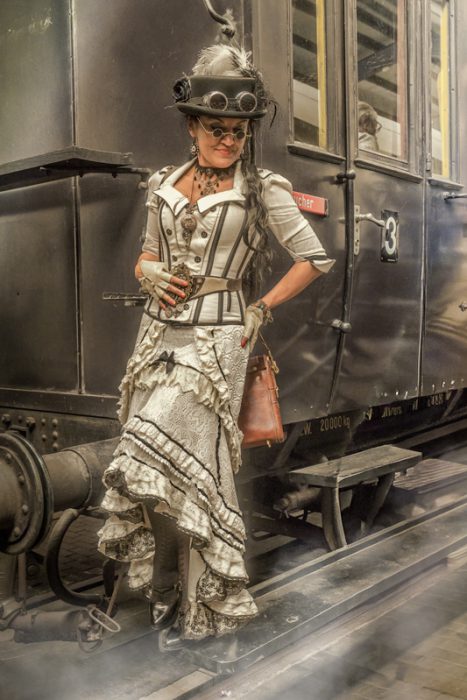 Eine Frau in einem weißen, korsettartigen Steampunk-Outfit mit Rüschenrock und Zylinderhut, hält eine kleine Vintage-Tasche und posiert vor einem dunkelgrünen historischen Zugwaggon.