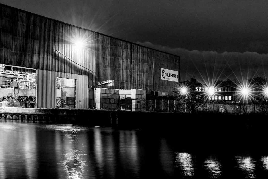 Monochrome Fotografie eines Hafengebiets bei Dunkelheit. Eine massive Lagerhalle am Wasserufer reflektiert helle Scheinwerfer. Im Hintergrund sind weitere Gebäude und Lichter erkennbar