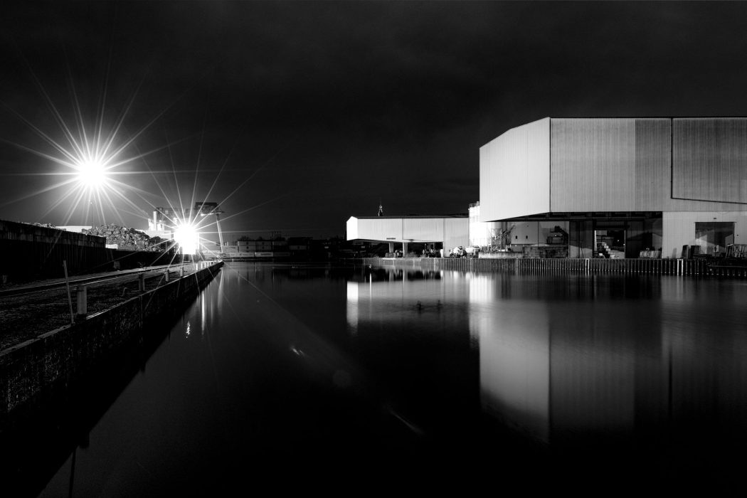 Schwarz-Weiß-Nachtansicht des Dortmunder Hafens. Moderne, quaderförmige Gebäude spiegeln sich in der ruhigen Wasseroberfläche. Links im Bild ein stark leuchtender Sternenstrahl von einem Licht.