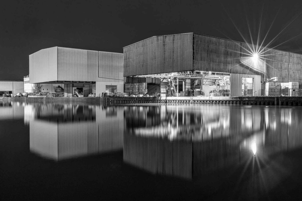 Schwarz-Weiß-Nachtansicht von zwei großen Industriehallen oder Lagerhäusern, die sich in einem dunklen Gewässer spiegeln. Helle Lichter, darunter ein sternförmiger Lichtstrahl, leuchten aus den Gebäuden
