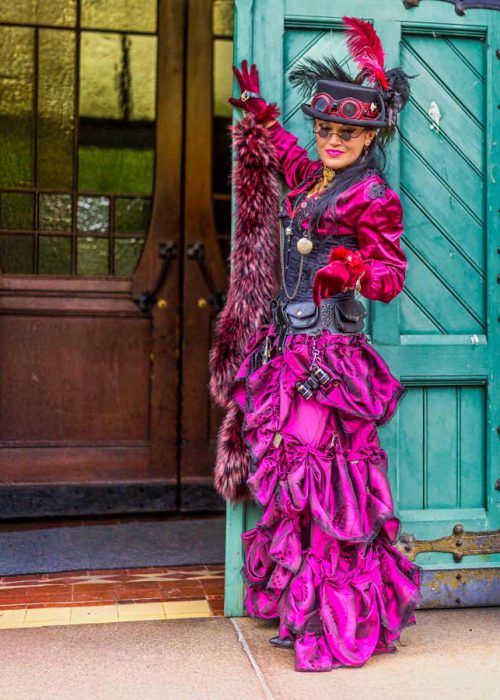 Eine lächelnde Frau in einem opulenten pinken und schwarzen Steampunk-Kostüm mit Rüschenrock, Korsett, Zylinder mit Federn und einer pelzigen Stola, steht in einem Türrahmen mit einer blauen Holztür im Hintergrund. Ihre Hand ist zum Gruß erhoben.