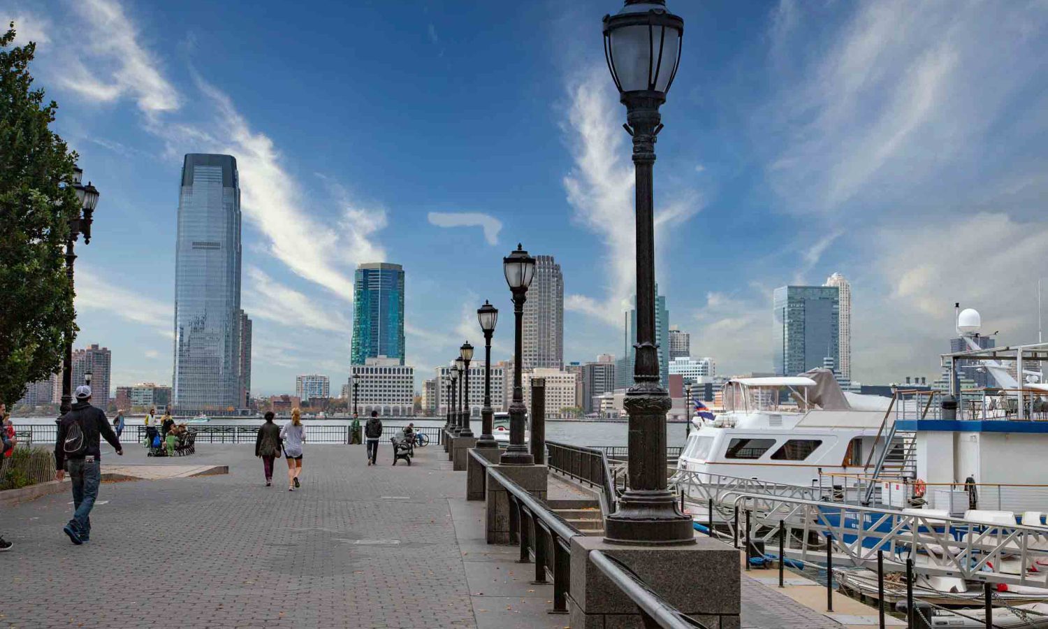 Dieses Bild zeigt die malerische Promenade des Battery Park in New York City an einem teilweise bewölkten Tag. Im Vordergrund erstreckt sich ein gepflasterter Weg mit klassischen Laternenmasten und einem Geländer, das zum Wasser hin abfällt. Entlang des Weges sind mehrere Personen zu sehen, die spazieren gehen oder die Aussicht genießen. Rechts sind Yachten und Boote an einem Pier vertäut. Im Hintergrund erhebt sich die imposante Skyline von Lower Manhattan oder Jersey City mit verschiedenen Hochhäusern und modernen Wolkenkratzern, die sich unter einem weiten blauen Himmel abzeichnen. Dieses Bild ist ideal für Suchanfragen zu New York City Sehenswürdigkeiten, Waterfront Parks, Stadtpromenaden, urbanen Landschaften und Reisefotografie.