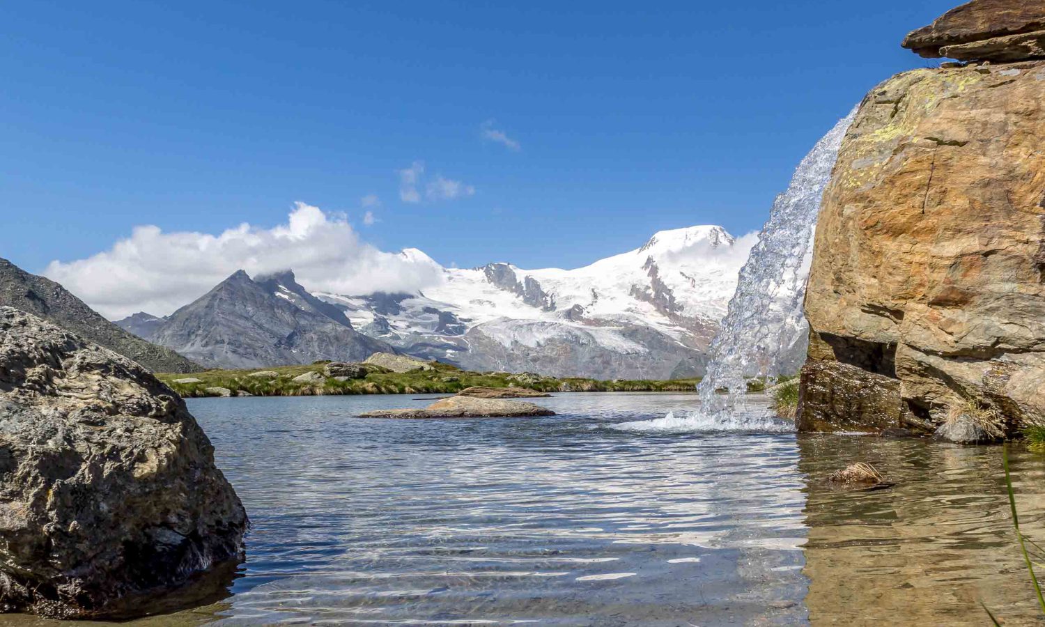 Bergsee mit kleinem Wasserfall über Felsen im Vordergrund, im Hintergrund schneebedeckte Alpen Gipfel mit Gletschern unter blauem Himmel – klare Berglandschaft im Sommer.
