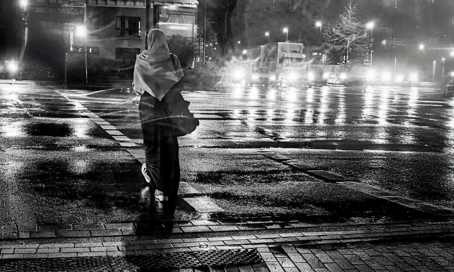 Schwarz-weiß Foto einer Frau mit Kopftuch, die bei Nacht und Regen an einer Straßenkreuzung wartet, Scheinwerfer von Autos spiegeln sich auf dem nassen Asphalt.
