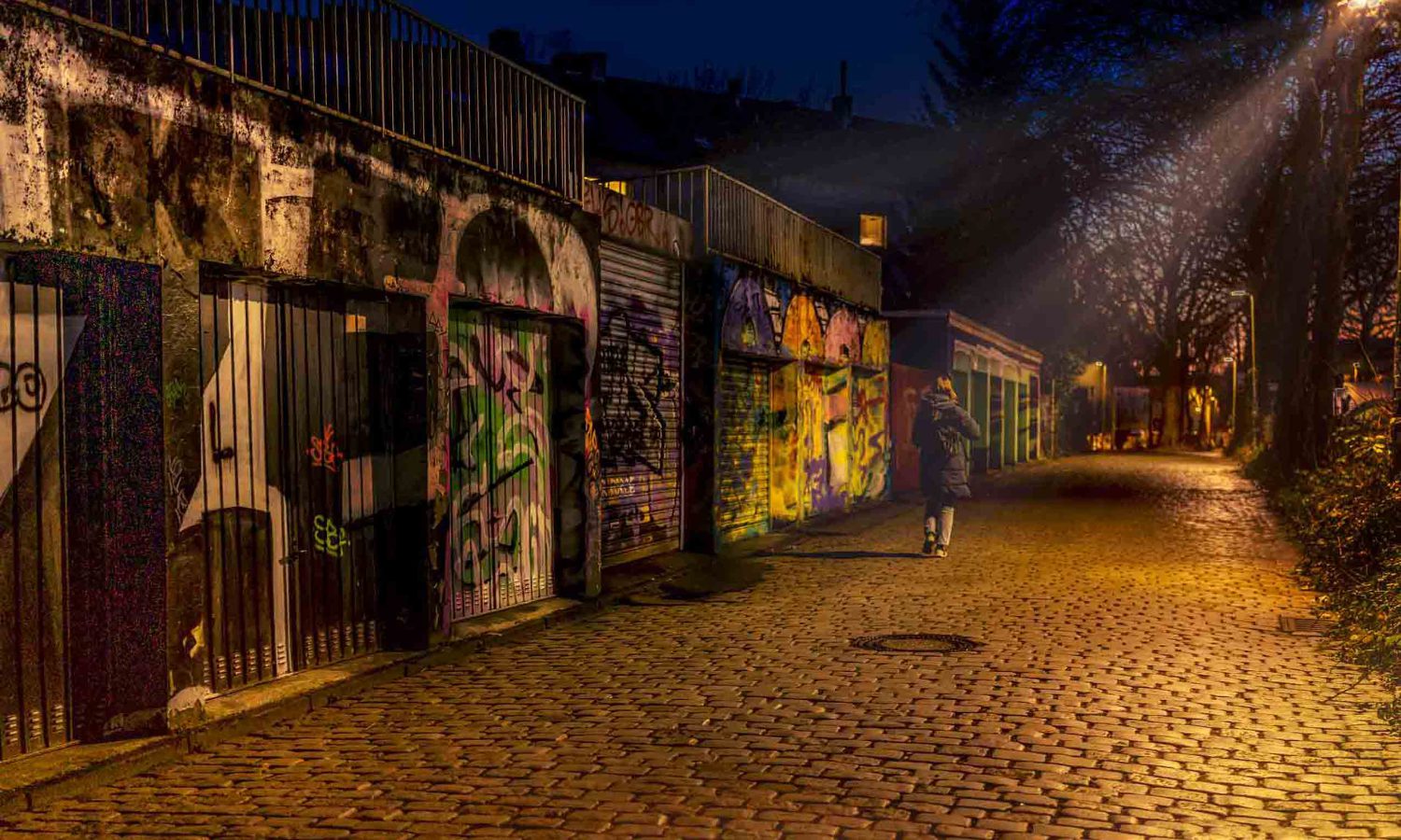 Person geht nachts allein über Kopfsteinpflasterweg an Garagen mit bunten Graffiti und Street Art vorbei, warmes Laternenlicht, Lichtstrahl durch kahle Bäume, urbane Atmosphäre bei blauer Stunde.