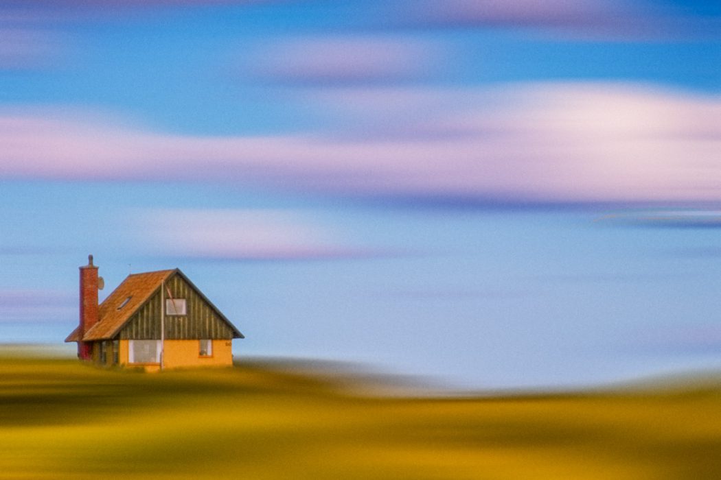 Eine künstlerische Fotografie eines kleinen gelben Hauses auf einem Hügel. Der Himmel und der Vordergrund sind durch horizontale Wischbewegungen in abstrakte blaue, violette und gelbe Farbbänder verwandelt.