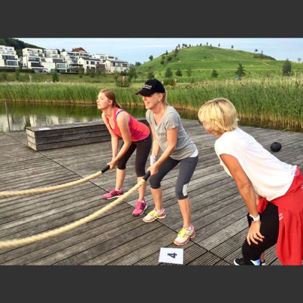 2 Frauen trainieren gemeinsam im Freien mit Battle Ropes auf einem Holzsteg an einem See. Im Vordergrund führt Petra Rimek als Fitness Coach das Seiltraining aus und Tipps gibt. Die Szene spielt in einer naturnahen Umgebung mit grüner Hügellandschaft, Schilf und moderner Wohnbebauung im Hintergrund. Alle Beteiligten tragen sportliche Outfits und wirken motiviert. Die Trainingseinheit findet bei Tageslicht statt und vermittelt ein Gefühl von Fitness, Gesundheit, Aktivität und Gemeinschaft im Freien.