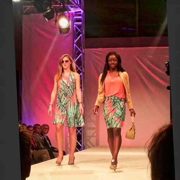 Modenschau-Laufsteg mit zwei Models: Zwei Models präsentieren farbenfrohe, gemusterte Outfits auf einem Laufsteg. Das linke Model trägt ein Kleid und Sonnenbrille, das rechte eine Kombination aus Rock und Oberteil mit einer beigen Jacke und Handtasche. Im Hintergrund sind Lichttechnik und Zuschauer angedeutet. Das Bild könnte eine Kollektion von Kimberly Goldson von Project Runway zeigen. Ideal für Mode-Events, Designer-Kollektionen und Fashion-Blogs.