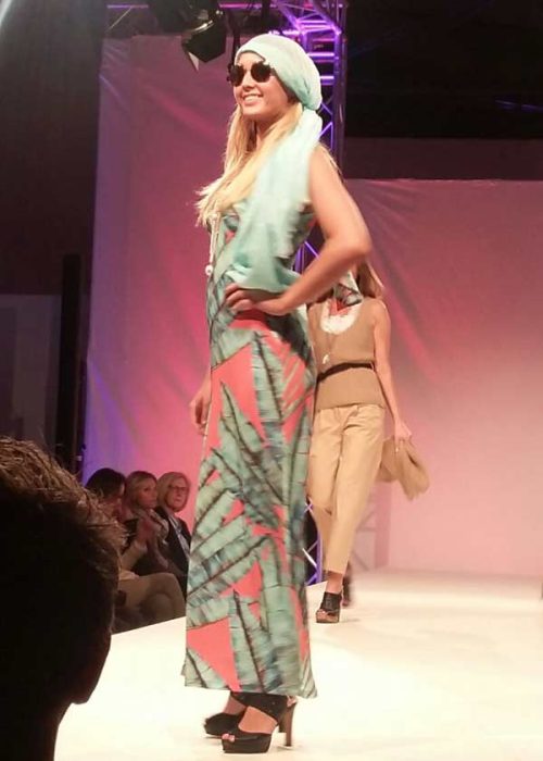 Ein lächelndes, blondes Model auf einem Laufsteg trägt ein langes, gemustertes Kleid in Türkis und Koralle, dazu ein passendes Kopftuch und Sonnenbrille. Eine weitere Person ist unscharf im Hintergrund zu sehen.