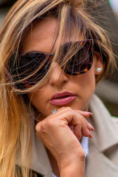 Nahaufnahme einer Frau mit Sonnenbrille, blond gesträhnten Haaren und beigem Trenchcoat.