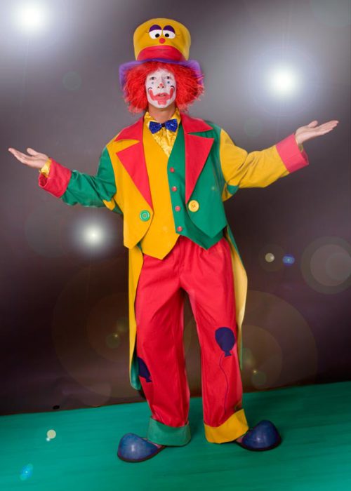 Ein Clown in einem farbenfrohen Kostüm aus gelb, rot und grün, mit einem hohen gelben Hut, roter Perücke und weißer Gesichtsbemalung, steht mit ausgebreiteten Armen und einem Lächeln auf einem grünen Boden. Im Hintergrund sind Lichtpunkte zu sehen.