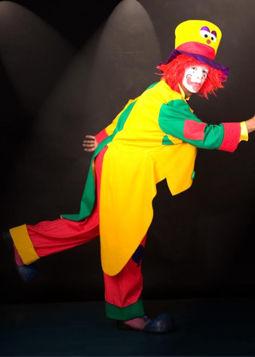 Ein Clown in einem leuchtend gelben, roten und grünen Kostüm mit großer gelber Melone, roter Perücke und weißer Gesichtsbemalung steht auf einem Bein und breitet die Arme aus. Zwei helle Spotlights sind im Hintergrund zu sehen.