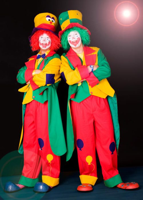 Zwei Clowns in leuchtend gelben, roten und grünen Kostümen stehen nebeneinander mit verschränkten Armen. Der linke Clown hat eine rote Perücke und einen gelben Hut, der rechte Clown eine grüne Perücke und einen grün-gelben Hut. Beide tragen weiße Gesichtsbemalung mit roten Wangen und Lippen.
