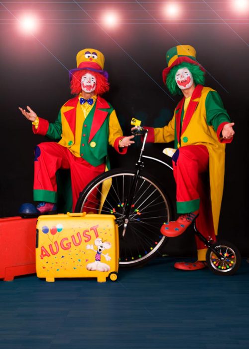 Zwei farbenfroh gekleidete Clowns posieren vor einem dunklen Hintergrund. Einer sitzt auf einem Hochrad, der andere steht daneben. Zwischen ihnen steht ein gelber Koffer mit der Aufschrift "AUGUST". Beide Clowns tragen hohe Hüte und expressive Schminke.