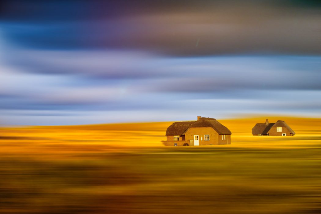 Künstlerische Fotografie von zwei Reetdachhäusern in einer abstrakt verschwommenen Landschaft. Der Vordergrund leuchtet in warmen Gelbtönen, während der Himmel in blauen und violetten Streifen verläuft.