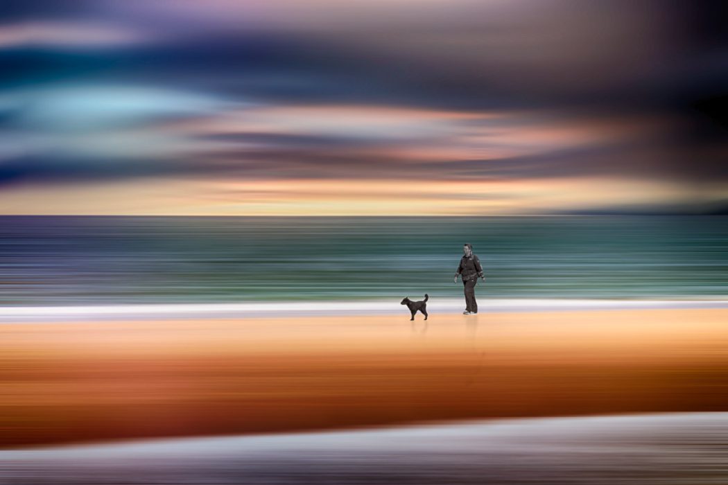Eine künstlerische Fotografie einer Person und eines kleinen Hundes an einem Strand. Durch horizontale Wischbewegungen verschwimmen die Umgebung und der Himmel in weiche Farbstreifen von Orange bis Dunkelblau, während die Silhouetten von Mensch und Tier klar erkennbar bleiben.