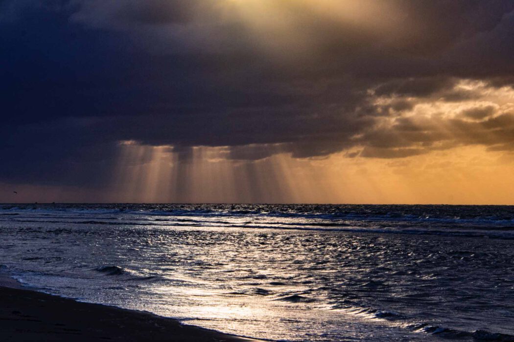 Reisefotografie Nordsee Strand bei Sturmlicht: Dunkle Wolken mit Sonnenstrahlen die durch Wolkenlücke brechen und auf Meer leuchten, goldene Reflexion auf Wellen, dunkler Sandstrand im Vordergrund, dramatisches Wetter.