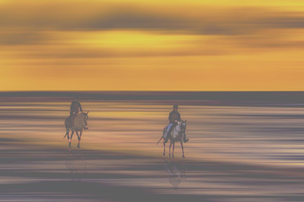 Zwei Reiter auf Pferden galoppieren über spiegelnden Sand bei Sonnenuntergang vor orange-violettem Himmel