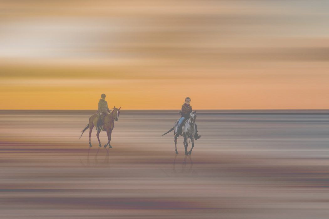 „Zwei Reiter auf Pferden am Strand bei Sonnenuntergang vor warmem Himmel mit Spiegelung im Sand