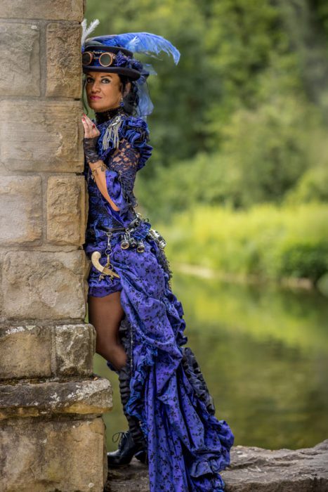 Frau in lila Steampunk-Kleid an Steinmauer.