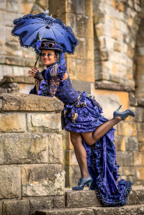 Frau in lila Steampunk-Kleid an Steinmauer.