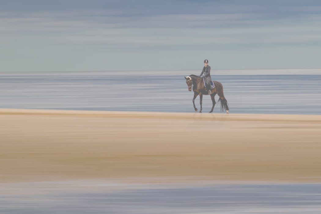 Reiter auf dunkelbraunem Pferd am Strand vor ruhigem Meer und horizontal verschwommenem Himmel in Blau und Weiß