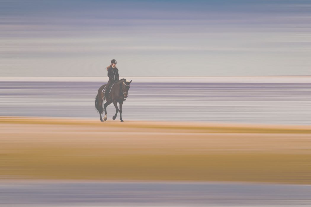 Reiter auf dunklem Pferd am Strand vor abstraktem, horizontal verschwommenem Hintergrund in Blau, Beige und Violett