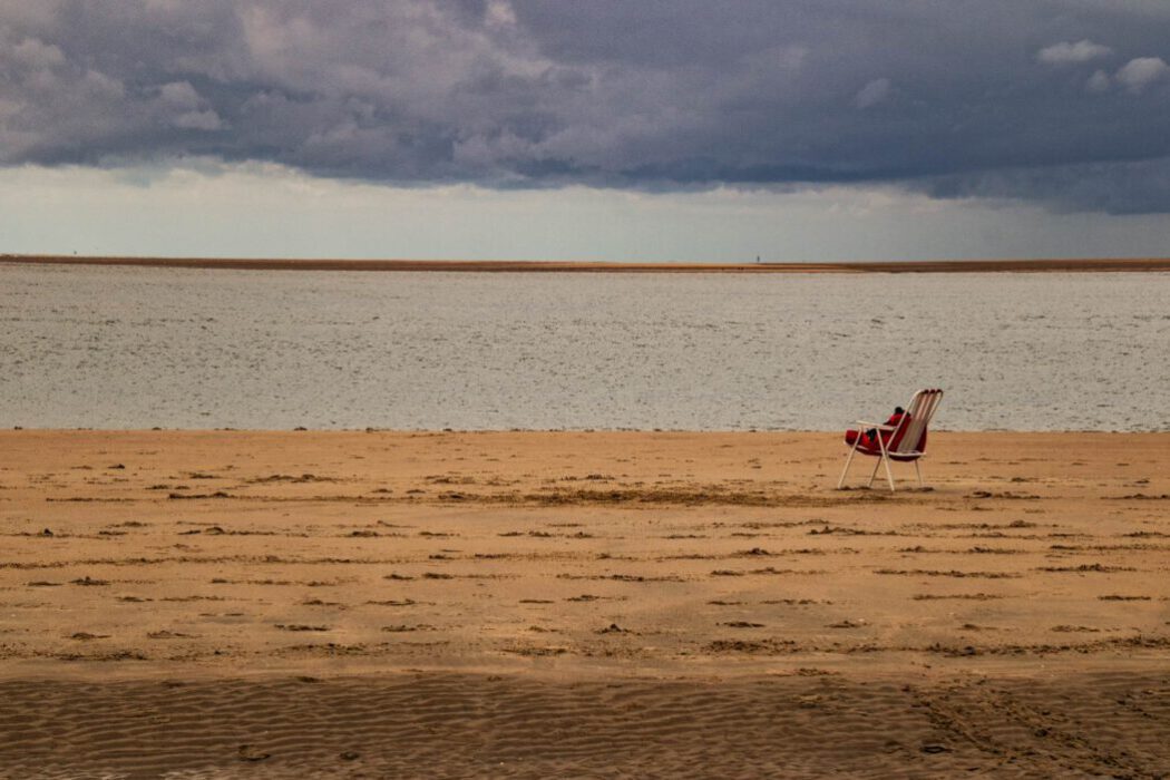 Ein leerer rot-weiß gestreifter Liegestuhl steht einsam am verlassenen Sandstrand unter dunklen Gewitterwolken und blickt auf eine weite Wasserfläche und eine Sandbank am Horizont.