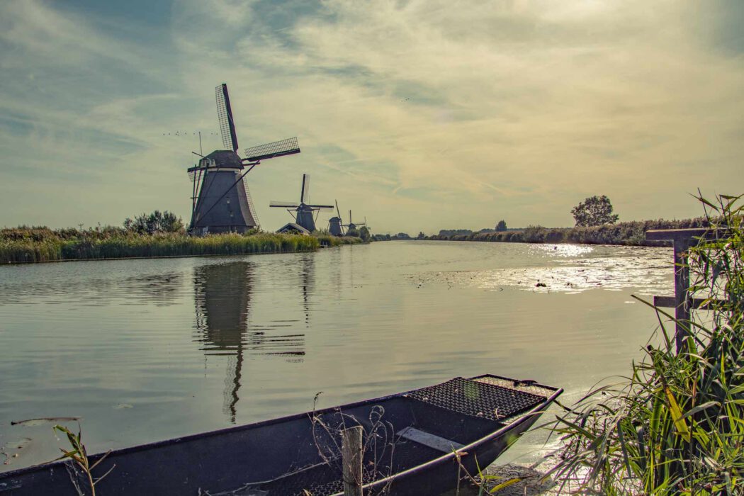 Historische Windmühlen entlang eines ruhenden Kanals mit Spiegelung im Wasser, im Vordergrund ein flaches Boot am Schilfufer unter leicht bewölktem Himmel.