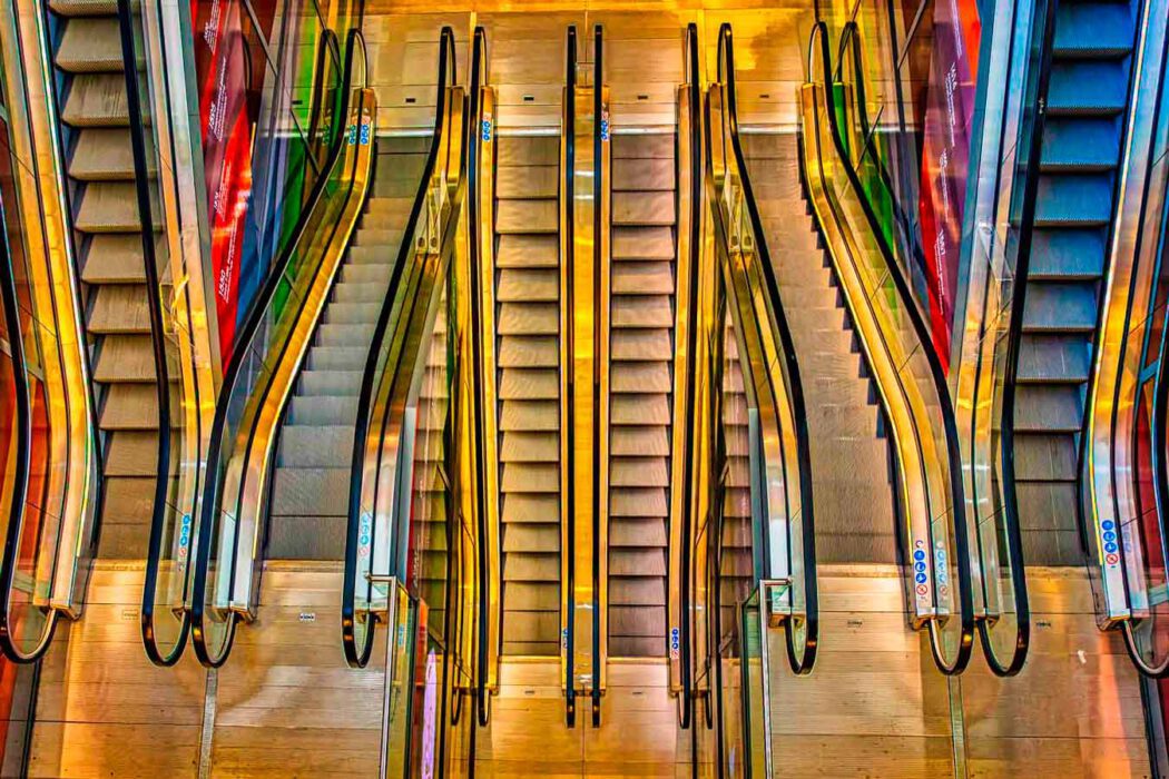 Symmetrische Draufsicht auf mehrere parallel verlaufende Rolltreppen mit leuchtend gelben Handläufen und farbenfrohen Reflexionen.