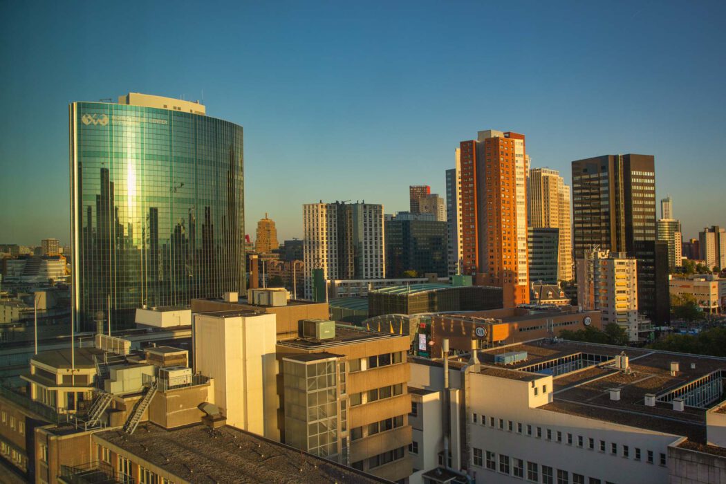 Reisefotografie Skyline Rotterdam im Abendlicht: World Trade Center Rotterdam mit grüner Glasfassade in der Sonne, dahinter Hochhäuser Zentrum, rote und braune Wohntürme, Dächer und Shopping Center Beursplein, goldenes Licht.