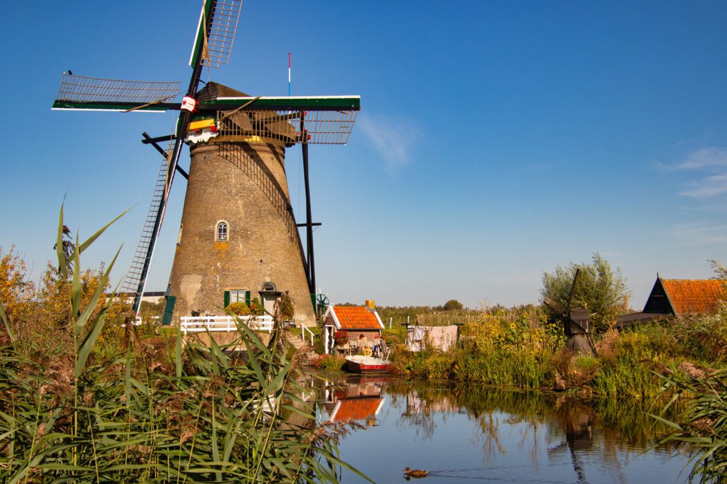 Historische Windmühle in einer niederländischen Landschaft mit Schilf, Wasserlauf und kleinen Reetdachhäusern unter blauem Himmel.