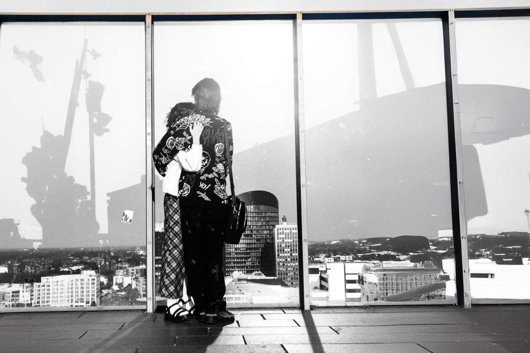 Schwarz-Weiß-Ansicht von zwei Personen, die von hinten vor einem großen Fenster stehen und auf eine Stadt mit Hochhäusern und reflektierten Schatten blicken