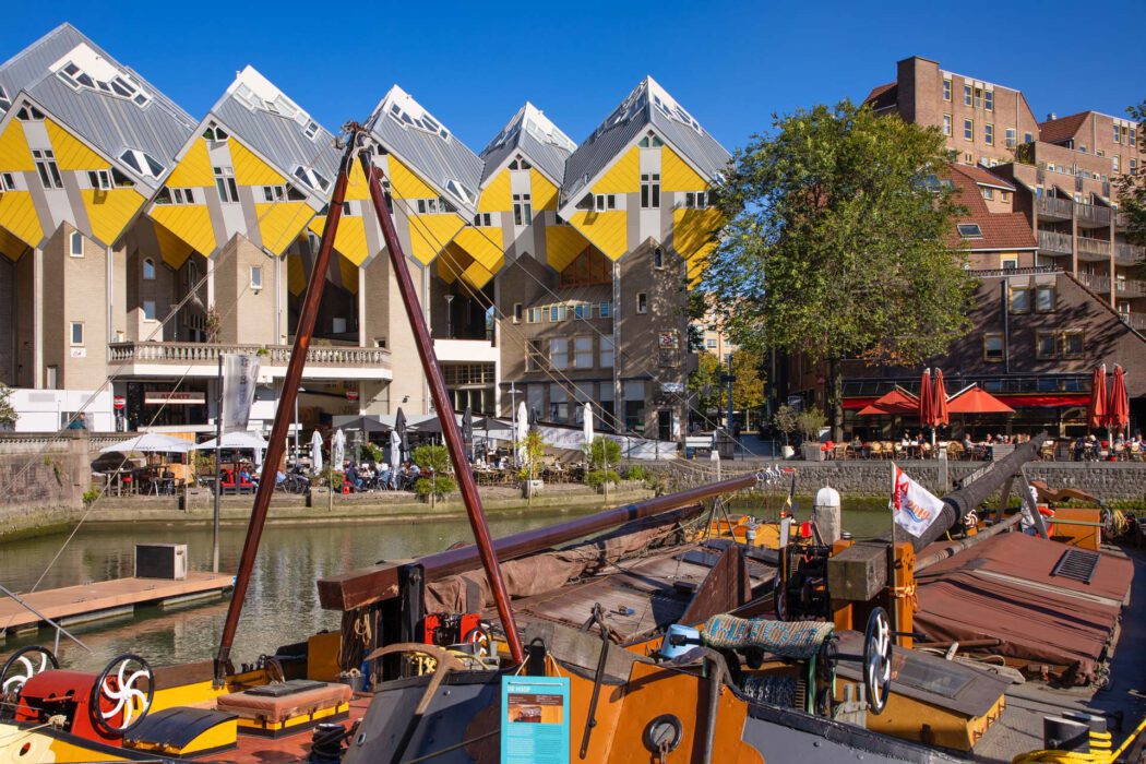 Kubushäuser in Rotterdam mit ihren markanten gelben, schräg geneigten Fassaden, aufgenommen am Wasser mit einem historischen Lastkahn im Vordergrund.