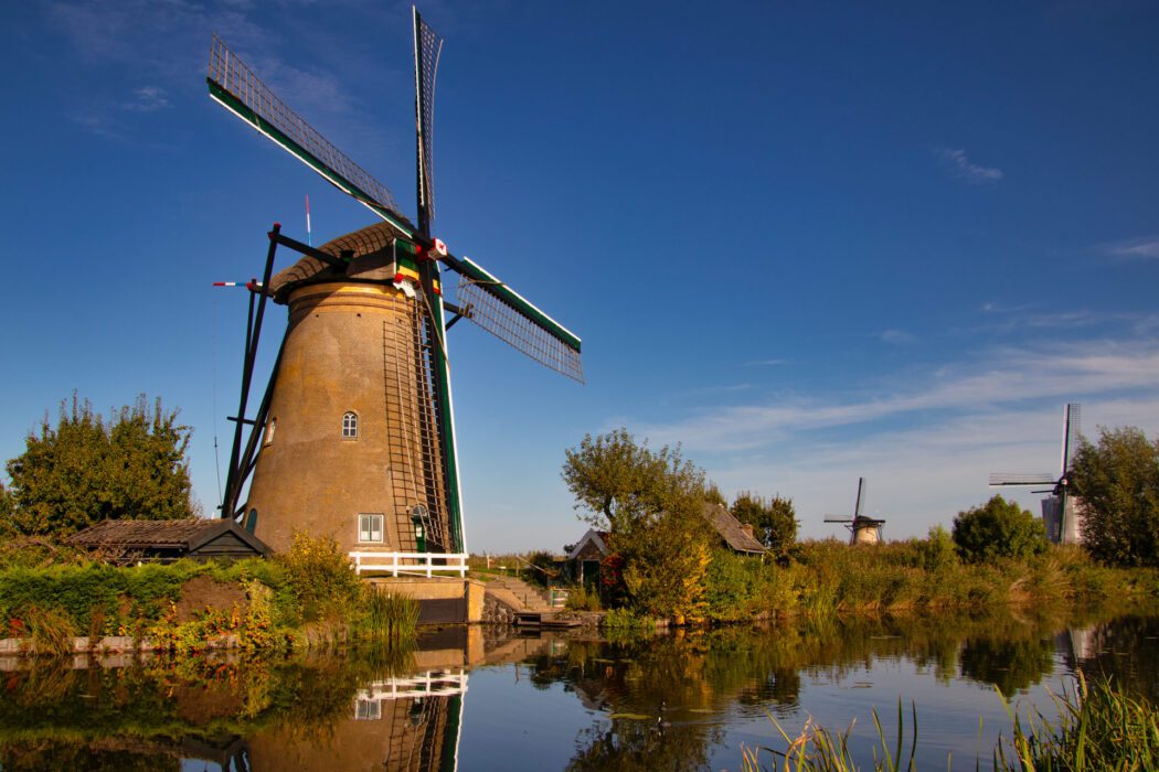 Ein großer traditioneller Windmühlenbau aus Backstein mit hölzernen Flügeln am Rand eines ruhigen Kanals; im Wasser spiegelt sich die Mühle, umgeben von grünen Pflanzen, einem kleinen Haus und weiteren Windmühlen im Hintergrund unter blauem Himmel.