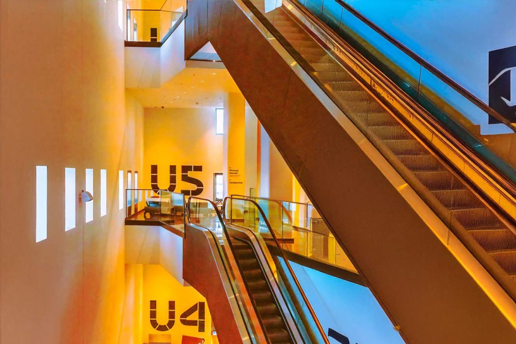 Innenansicht eines modernen Gebäudes mit zwei Rolltreppen, die sich über mehrere Etagen erstrecken. Die Wände sind hellgelb beleuchtet und zeigen die Beschriftungen 'U4' und 'U5'."