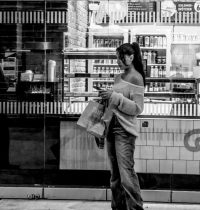 Junge Frau mit Pferdeschwanz in schulterfreiem Pullover und Jeans steht mit Einkaufstüte vor Bäckerei Schaufenster, Schwarz-Weiß Street Fotografie, Spiegelungen im Glas.
