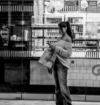 Eine Frau steht vor einem beleuchteten Kiosk, schaut auf ihr Smartphone und hält eine Einkaufstüte in der Hand.