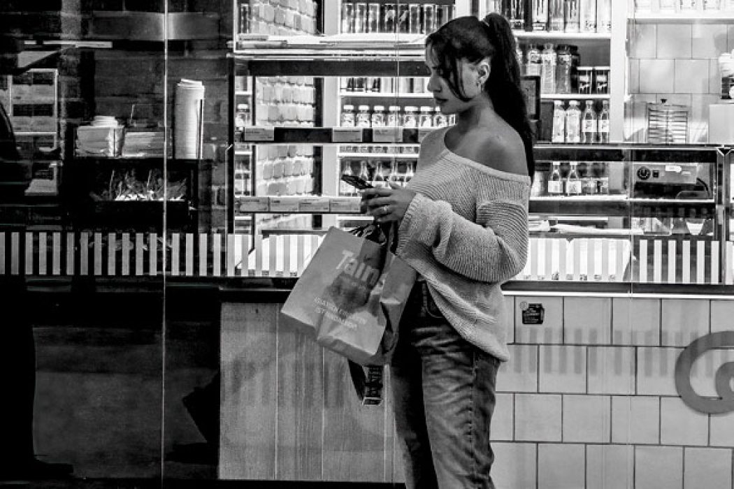 Eine Frau steht vor einem beleuchteten Kiosk, schaut auf ihr Smartphone und hält eine Einkaufstüte in der Hand.