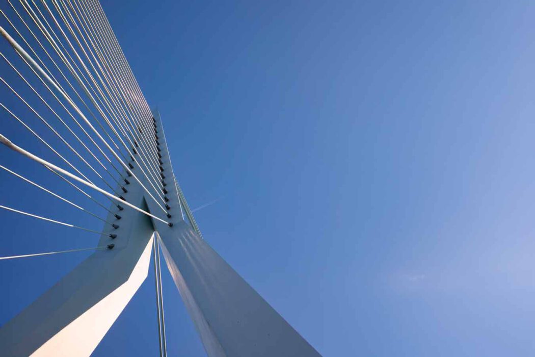 Ein schräg nach oben gerichteter Blick auf einen hellen Brückenpylon, von dem zahlreiche Stahlseile fächerförmig in den klaren blauen Himmel ziehen; die Linien wirken präzise, geometrisch und spannungsvoll.
