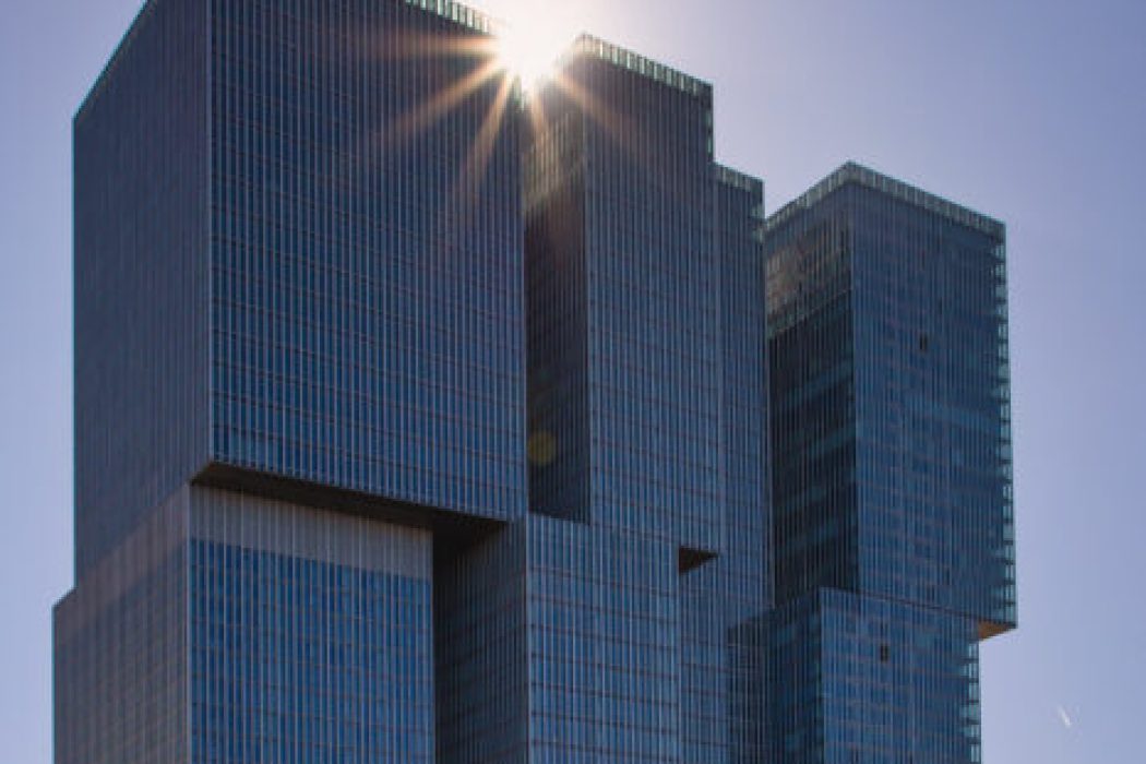 Drei hohe, gläserne Türme mit streng gerasterter Fassade; hinter dem obersten bricht die Sonne hervor und erzeugt einen strahlenden Lichtstern, während ein Flugzeug mit Kondensstreifen über den klaren Himmel zieht.