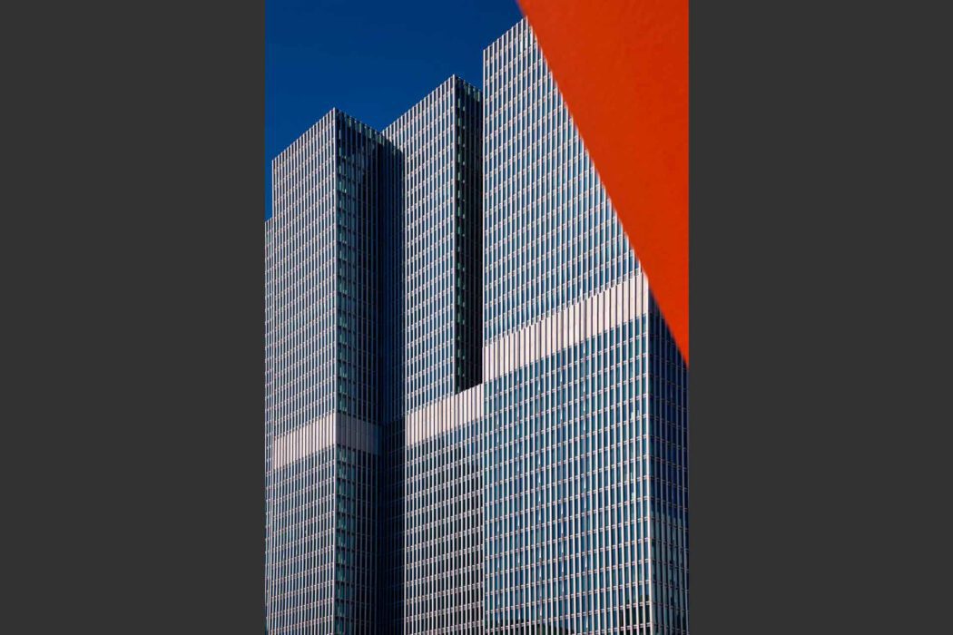 Reisefotografie De Rotterdam von OMA Rem Koolhaas: Drei gestapelte Glastürme mit vertikaler Lamellenfassade vor tiefblauem Himmel, im Vordergrund angeschnittene rote Stahlskulptur The Destroyed City, grafische Architekturkomposition.