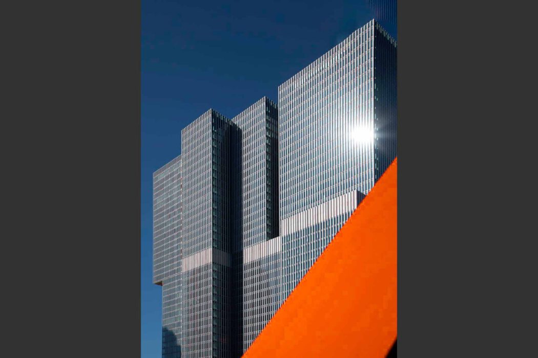 Reisefotografie De Rotterdam von OMA Rem Koolhaas: Drei gestapelte Glastürme mit vertikaler Lamellenfassade vor tiefblauem Himmel, im Vordergrund angeschnittene rote Stahlskulptur The Destroyed City, grafische Architekturkomposition.